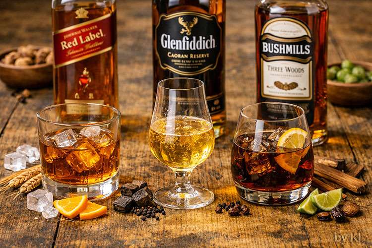 Whiskys Drei Whiskys stehen nebeneinander auf einem rustikalen Holztisch, jeder in einem passenden Glas mit eigener Farbe und Charakter:</p>
<p>Johnnie Walker Red Label: Ein klassischer Tumbler mit dickem Boden, gefüllt mit goldenem Whisky und Eiswürfeln. Das Getränk wirkt klar und lebendig, mit leichtem Rauchduft und würziger Note.</p>
<p>Glenfiddich Caoran Reserve: Ein Glencairn‑Glas mit goldgelbem Whisky, leicht ölig und aromatisch. Der Duft ist warm, mit Anklängen von Torf und Malz.</p>
<p>Bushmills Three Woods: Ein rundes Snifter‑Glas mit tief bernsteinfarbenem Whisky, leicht viskos, mit Noten von Holz und Trockenfrüchten.</p>
<p>Im Hintergrund stehen die dazugehörigen Flaschen, deren Etiketten farblich abgestimmt sind: Rot, Schwarz und Creme. Auf dem Tisch liegen Eiswürfel, Orangenscheiben, Hopfen, Holzstücke und Rosinen als Dekoration.</p>
<p>Das Licht ist warm und natürlich, die Stimmung ruhig, edel und authentisch – ideal für eine Whisky‑Verkostung oder eine hochwertige Getränkekarte.